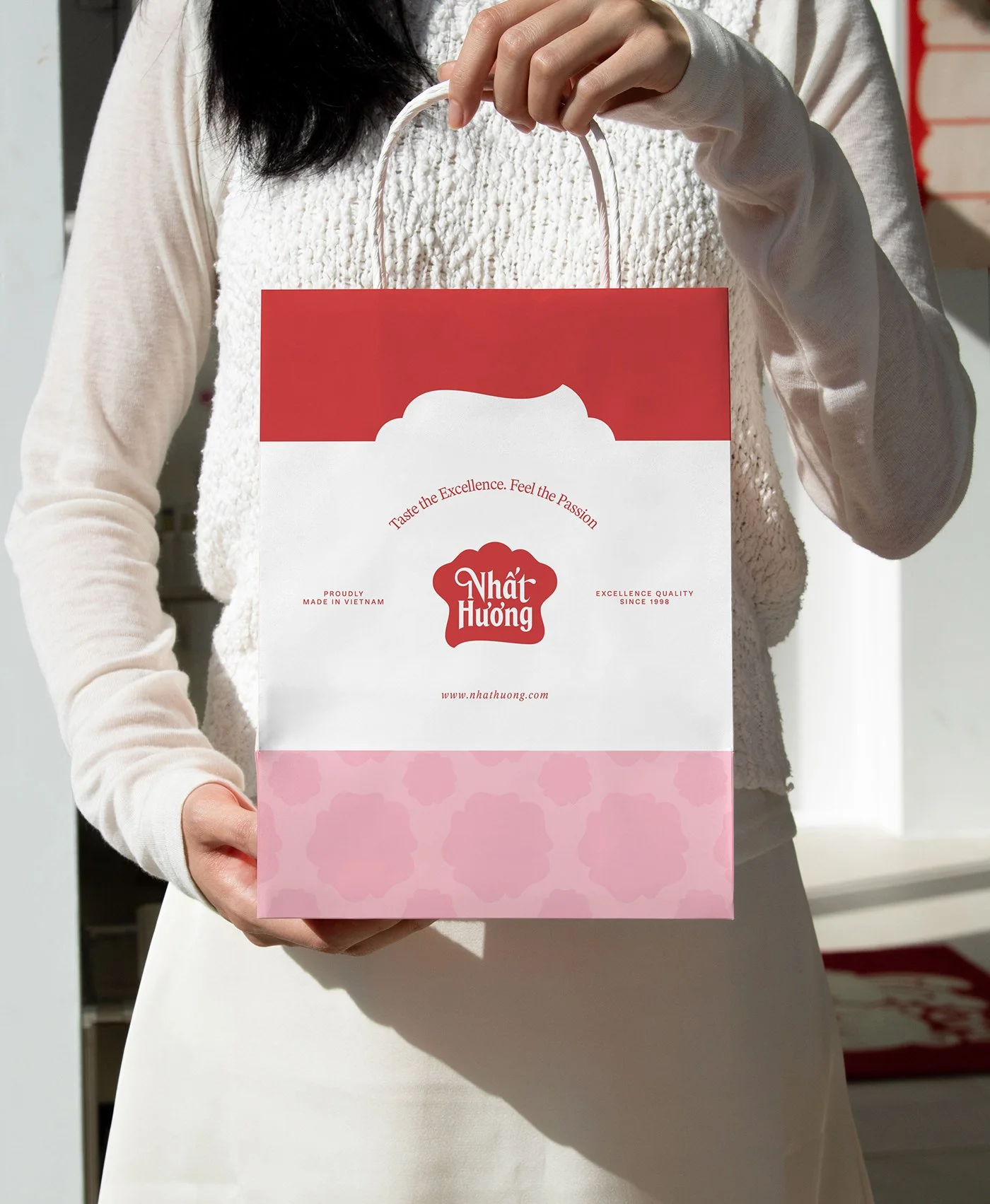 A person holding the Nhat Huong paper shopping bag: red top, white middle with the hat logo, candy-pink patterned base
